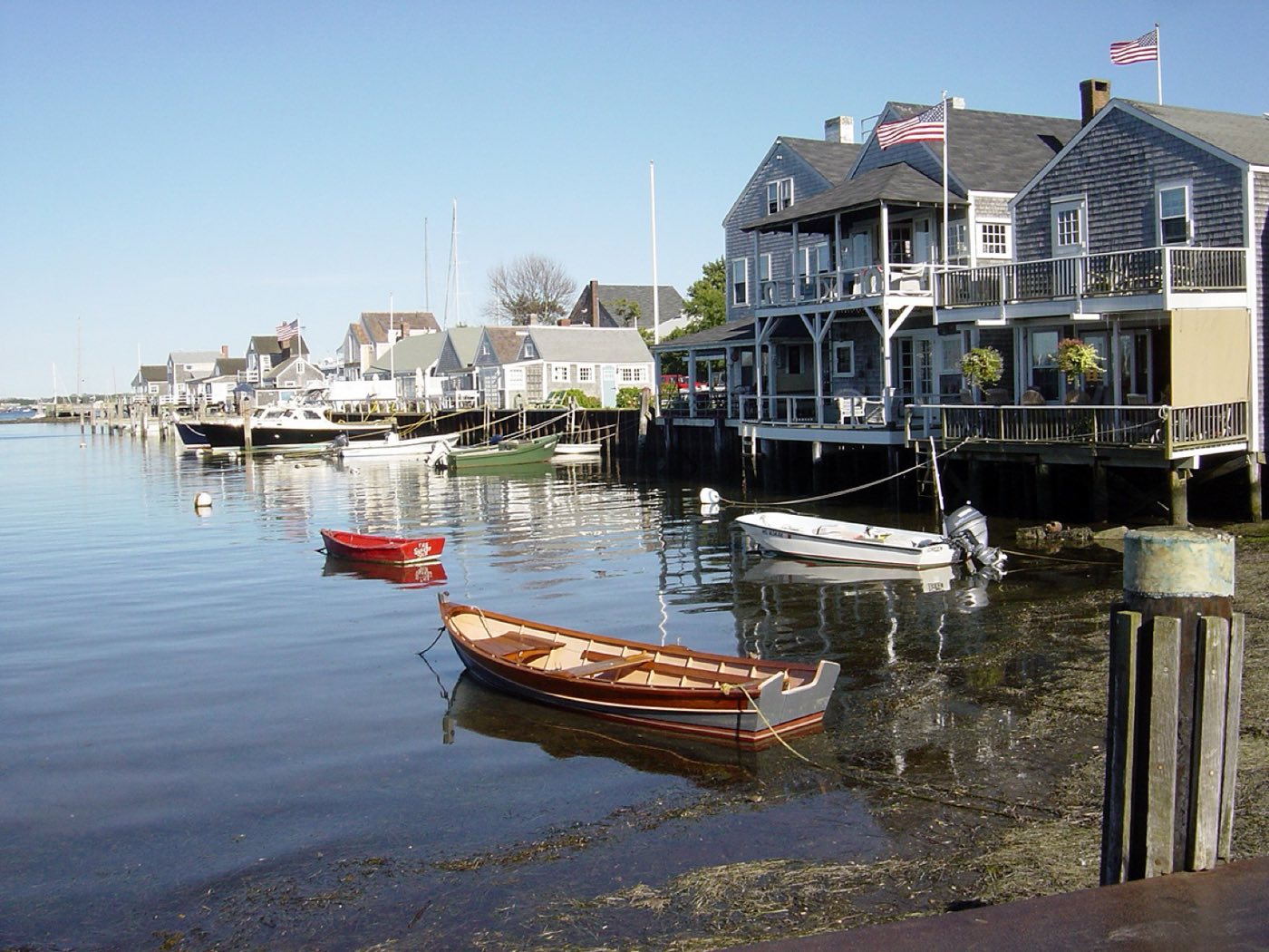 Small boats moored along a wharf of gray-shingled, flag-flying buildings on Nantucket Harbor.